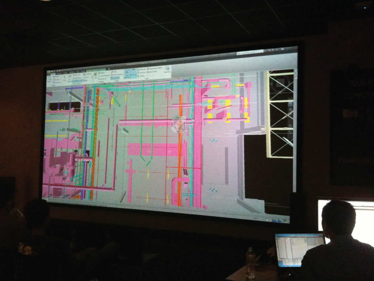 Photograph of a man in a dark room seated in front of a screen displaying the pipeline model for a project.