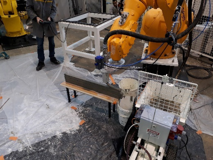 A photograph of a machine involved in the process of 3D printing mortar through extrusion and filament deposition, utilizing a pump and a six-axis robotic arm. The image appears to be captured outdoors, possibly showcasing the technology in action. 