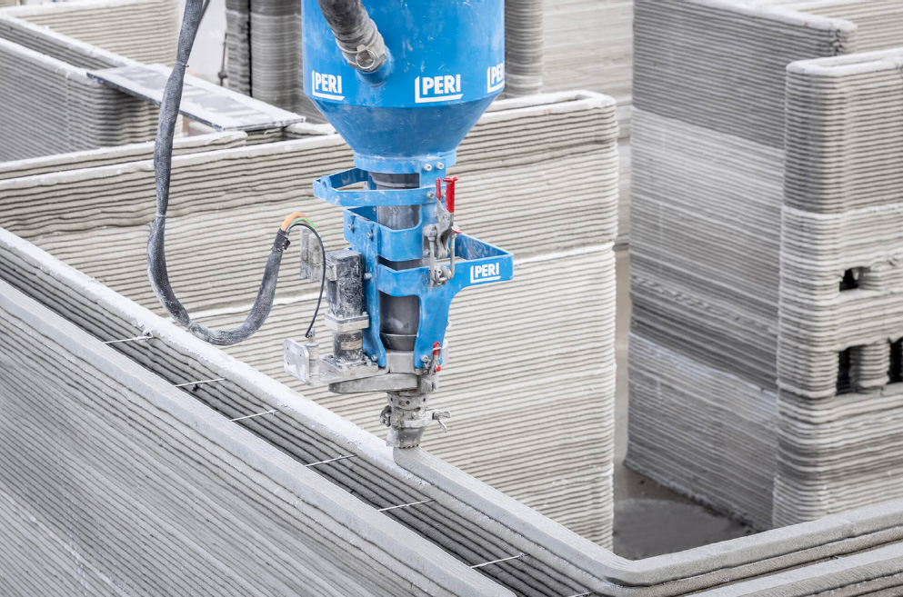 An image shows a close-up of a 3D-printed wall being constructed. A large, blue, robotic arm is extruding concrete to build the wall. The arm appears to be depositing concrete into a series of metal spacers, which are embedded in the wall.