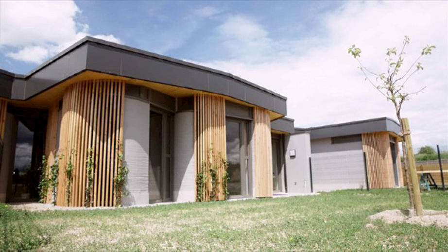 An image features a house with a grass yard, showcasing the south facade as part of the Villiaprint project. The scene includes elements such as a clear sky, clouds, trees, and greenery, highlighting aspects of outdoor living and real estate.