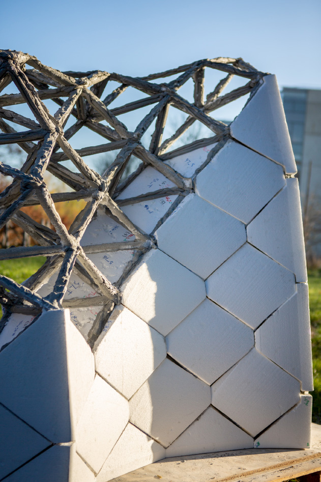 An image features a large metal structure, specifically a curved concrete truss. It was presented at the “F O R M and F O R C E” international event. The setting is outdoors with a clear view of the sky.